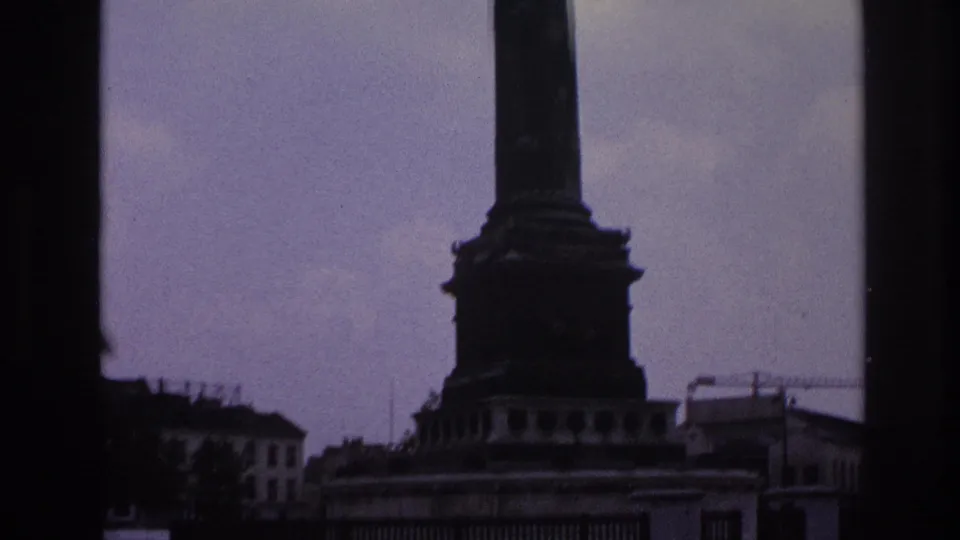1967 Paris home-movie frame showing a monument base through dark window edges with mid-century cars moving across the street.