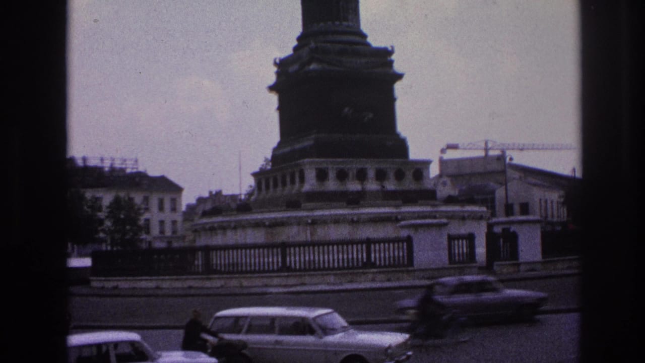 1967 Paris street frame with classic cars and a motorcyclist moving past a prominent monument base and perimeter railing.