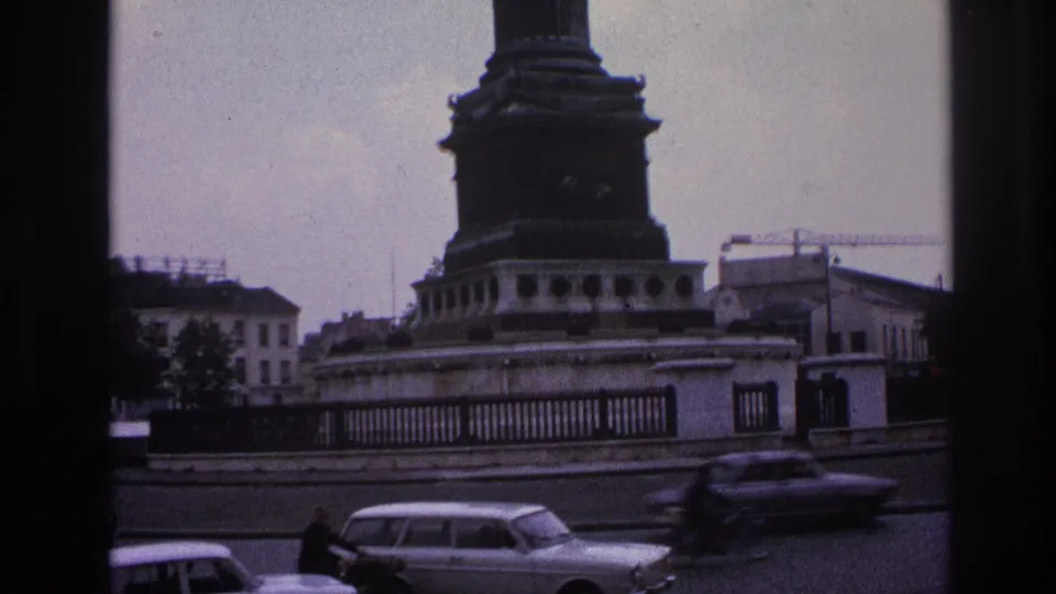 1967 Paris street frame with classic cars and a motorcyclist moving past a prominent monument base and perimeter railing.