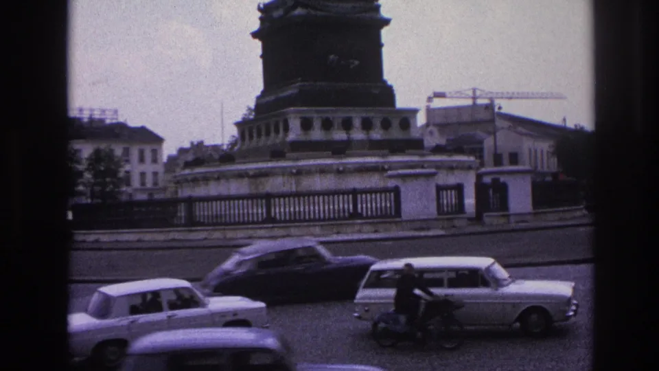 1967 Paris home-movie frame showing classic cars and a motorcyclist crossing the foreground near the base of a large monument.