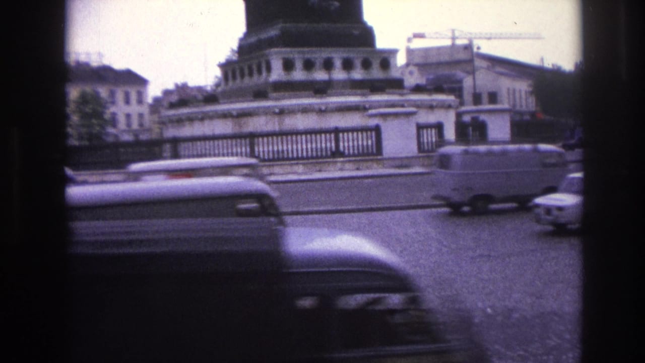 1967 Paris home-movie frame showing cars and a van moving around a roundabout beside stone architecture and a dark column base.