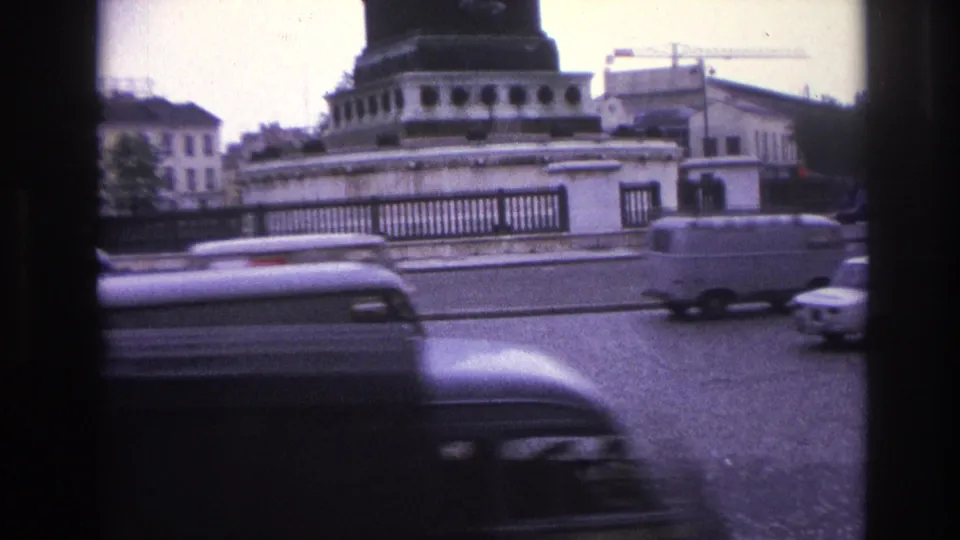 1967 Paris home-movie frame showing cars and a van moving around a roundabout beside stone architecture and a dark column base.