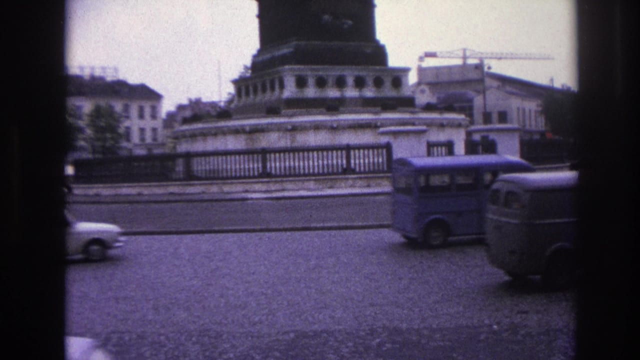 A blue van and other vehicles travel past a stone roundabout barrier.