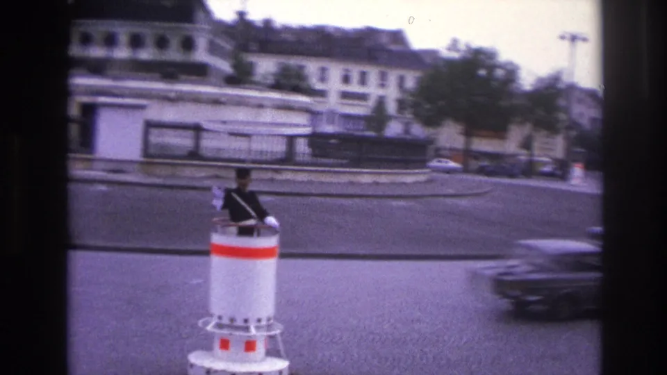 1967 Paris home-movie frame with a traffic officer on a red-and-white post, passing cars, stone barriers, trees, and city buildings.