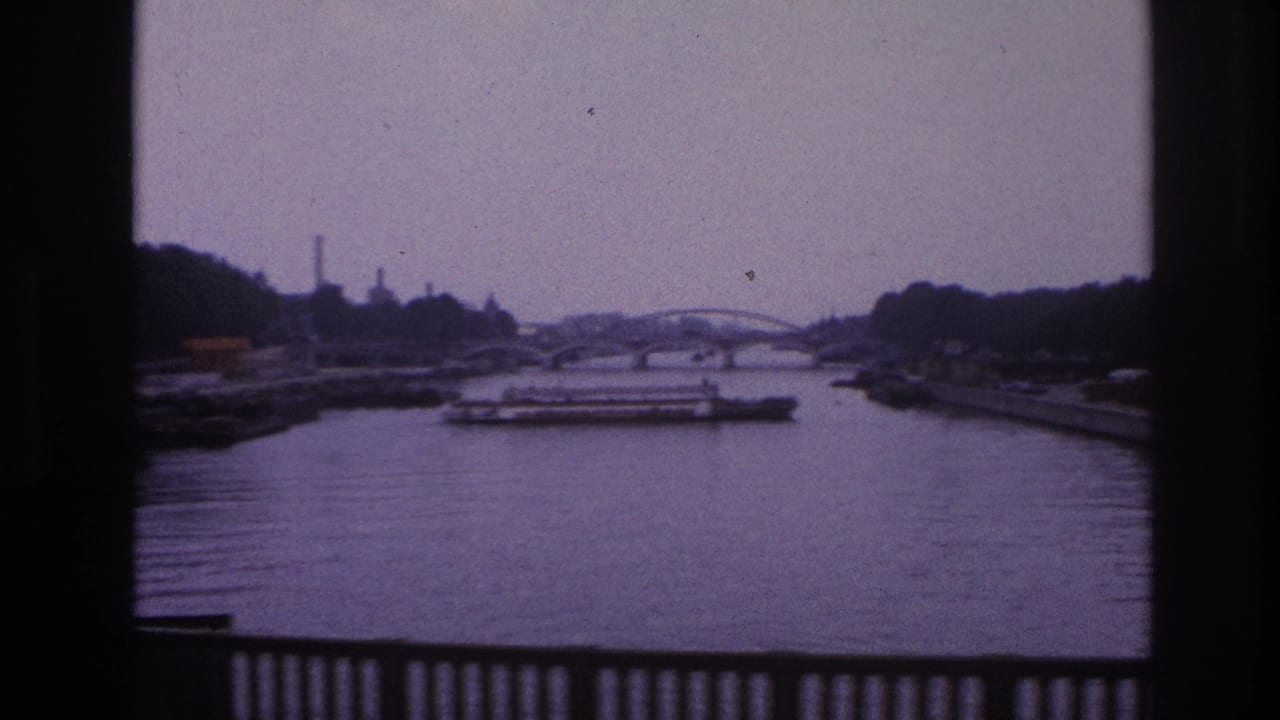 1967 Paris home-movie view of a river with a low freight barge, distant arched bridge, industrial background, and railing in the foreground.