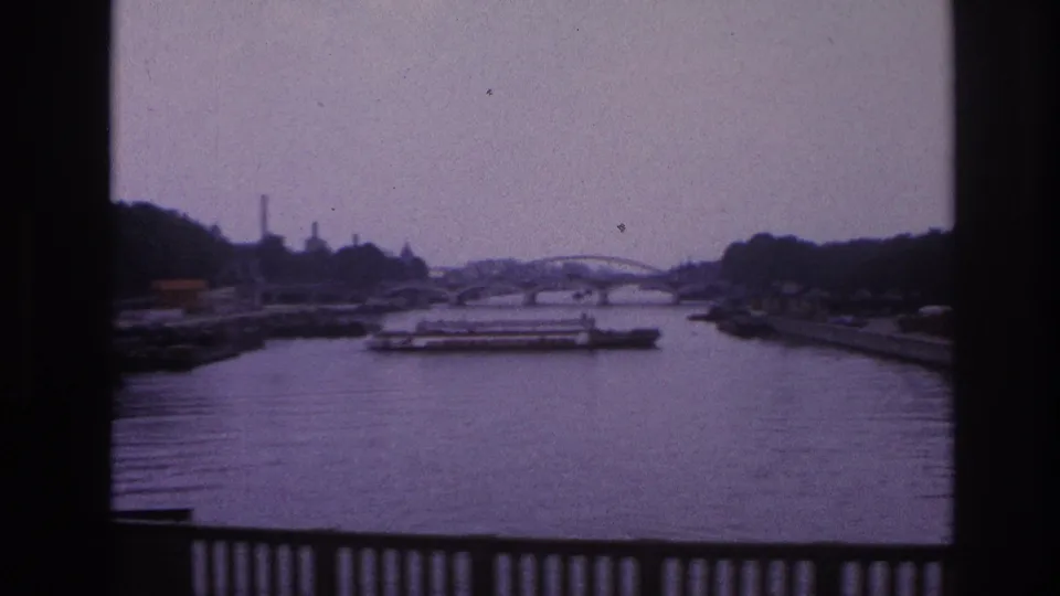 1967 Paris home-movie view of a river with a low freight barge, distant arched bridge, industrial background, and railing in the foreground.