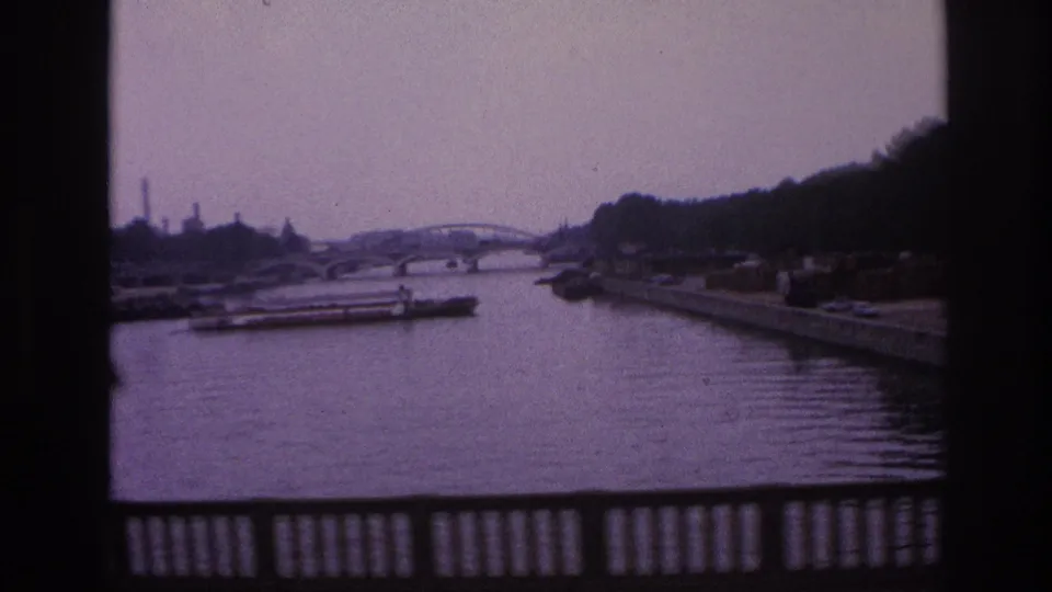 Soft 8mm footage from Paris in 1967 frames the river, a freight barge, an arched bridge, wooded bank, and dark foreground railing.