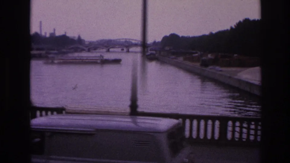 Final 1967 Paris frame shows the river and distant arched bridge beyond the railing, with a vehicle roof and vertical pole entering the foreground.