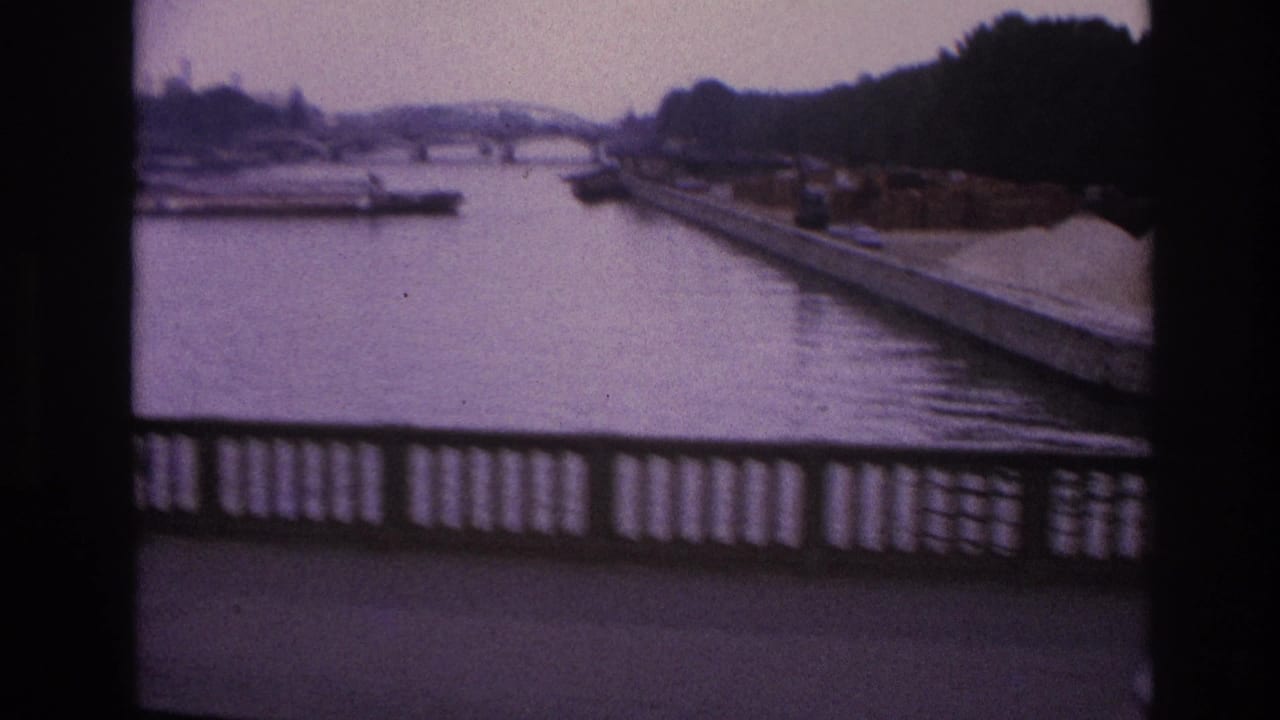 1967 Paris home-movie frame showing a river or canal view from above, with bridge railing, embankment, and dark bus-window framing.