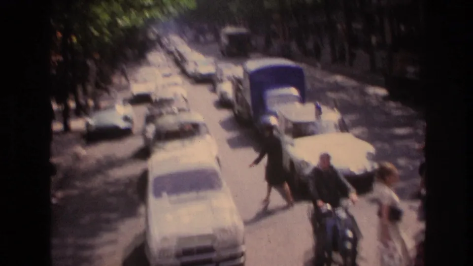 Upper-deck view of a 1967 Paris boulevard with cars, pedestrians crossing, and a scooter rider moving through traffic.