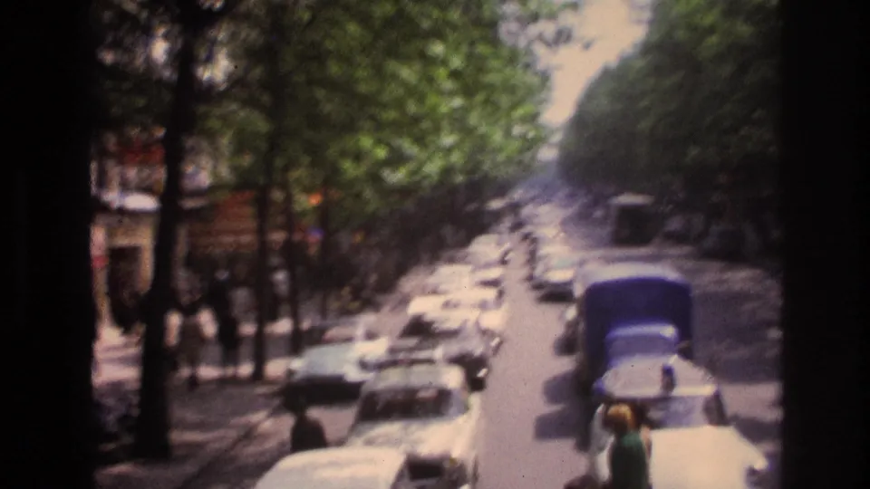 1967 Paris boulevard seen from a moving sightseeing bus, with rows of cars, pedestrians at the curb, and heavy tree cover along the street.