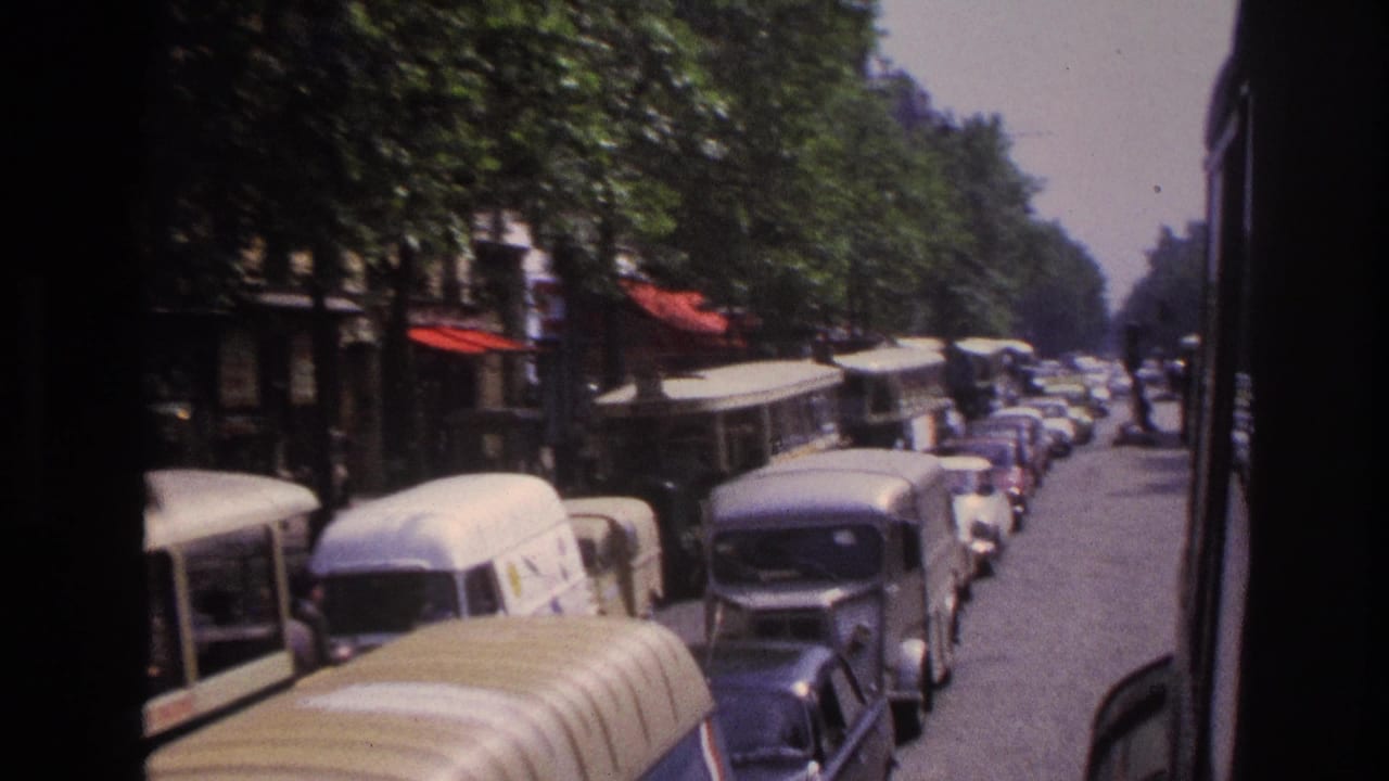 1967 Paris street view from a moving vehicle, showing older buses, dense parked cars, trees, and red storefront awnings.