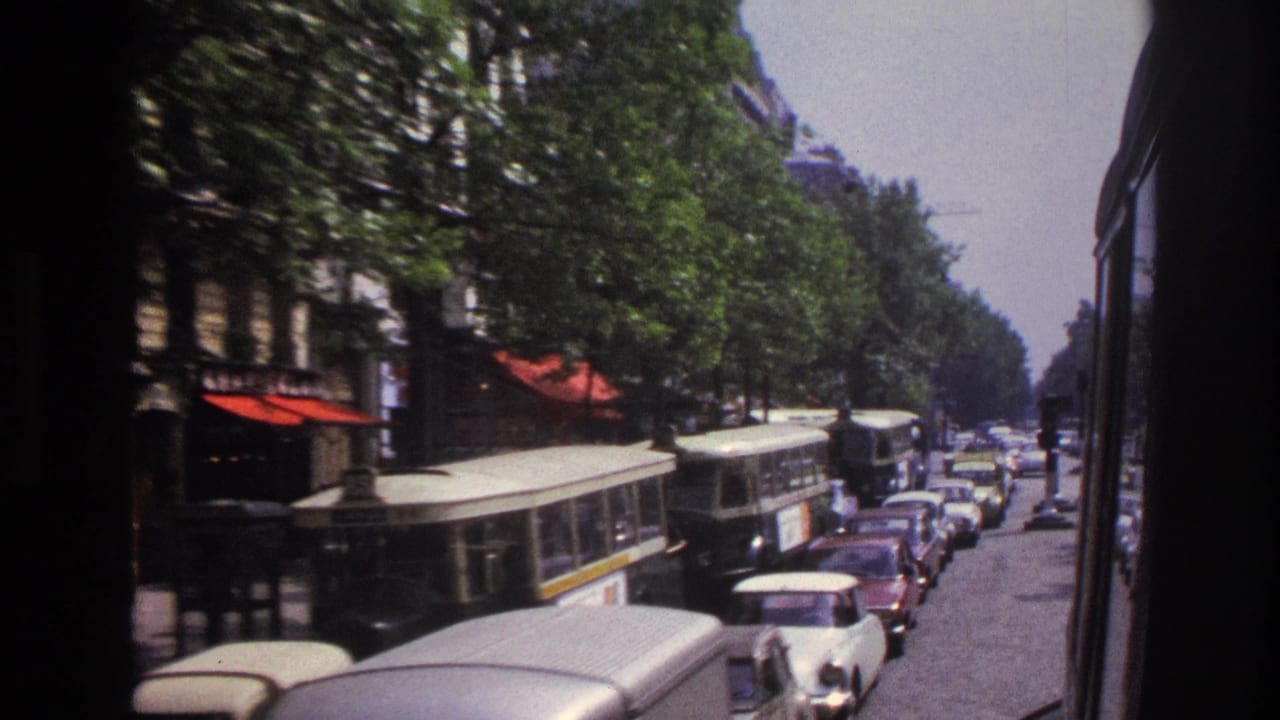 Street traffic with buses, compact cars, trees, storefronts, and a dark vehicle frame at the edge.