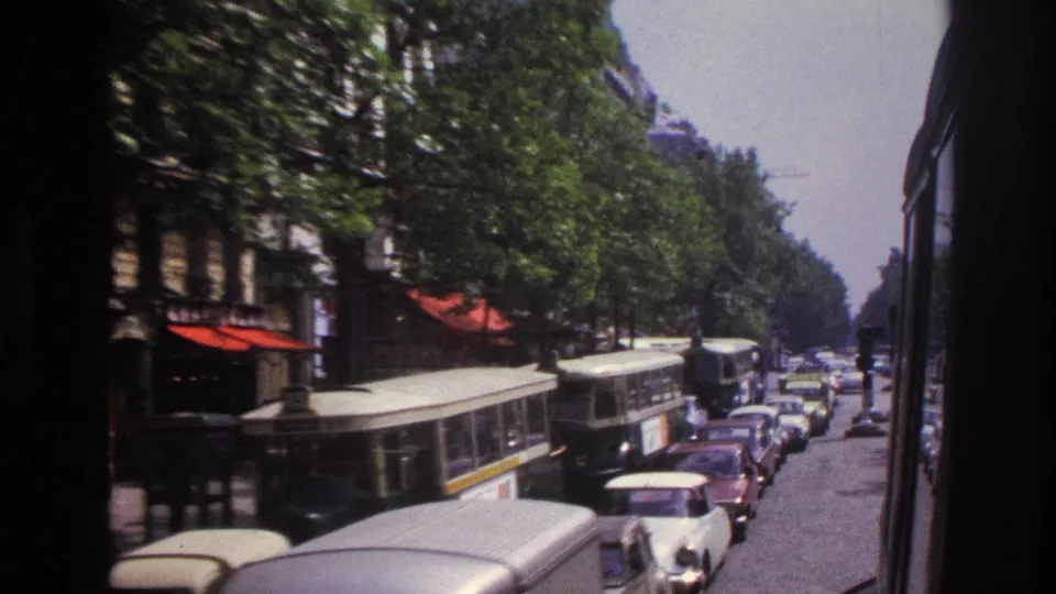 Moving 1967 Paris street perspective with cream-and-green buses, compact cars, trees, and red awnings along the sidewalk.