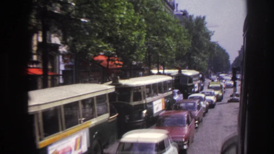 1967 Paris traffic scene with buses close to the camera, parked cars, shop awnings, and leafy trees along the avenue.