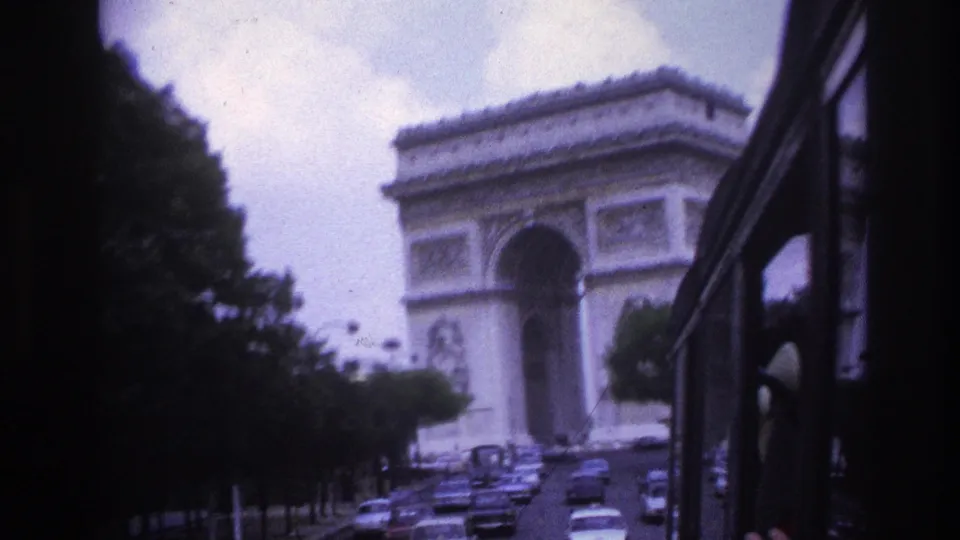 1967 Paris home-movie frame showing the Arc de Triomphe ahead through moving traffic and a vehicle window edge.