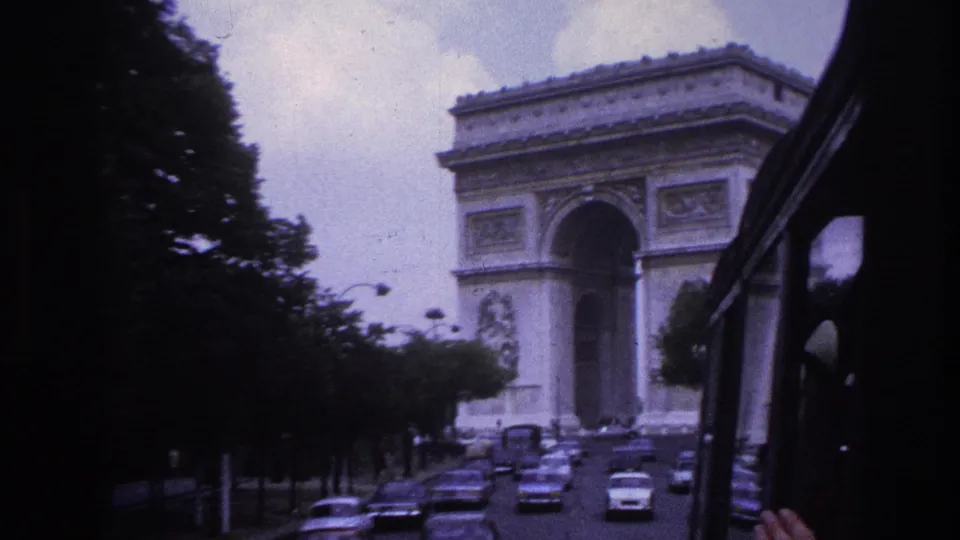 1967 Paris view of the Arc de Triomphe with mid-century traffic, filmed from a vehicle along a tree-lined avenue.