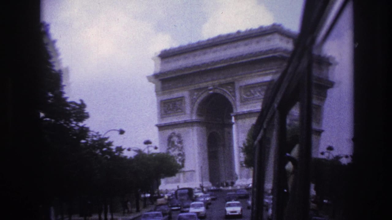 1967 Paris home-movie frame showing the Arc de Triomphe ahead through a vehicle window, with cars moving along the avenue.