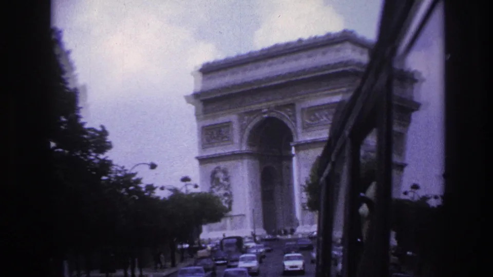 1967 Paris home-movie frame showing the Arc de Triomphe ahead through a vehicle window, with cars moving along the avenue.
