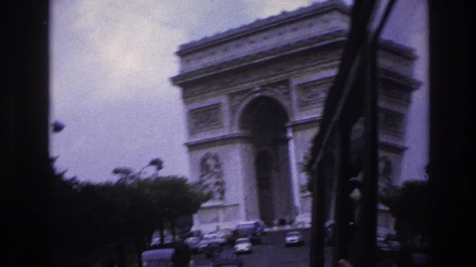 A tilted passenger view in Paris shows the Arc de Triomphe, roadside trees, and passing traffic through the vehicle window frame.