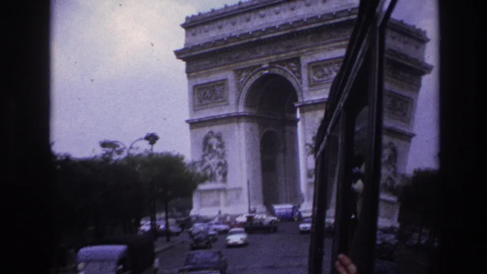 1967 Paris home-movie frame showing the Arc de Triomphe from inside a moving vehicle, with cars, trees, and a dark window edge visible.