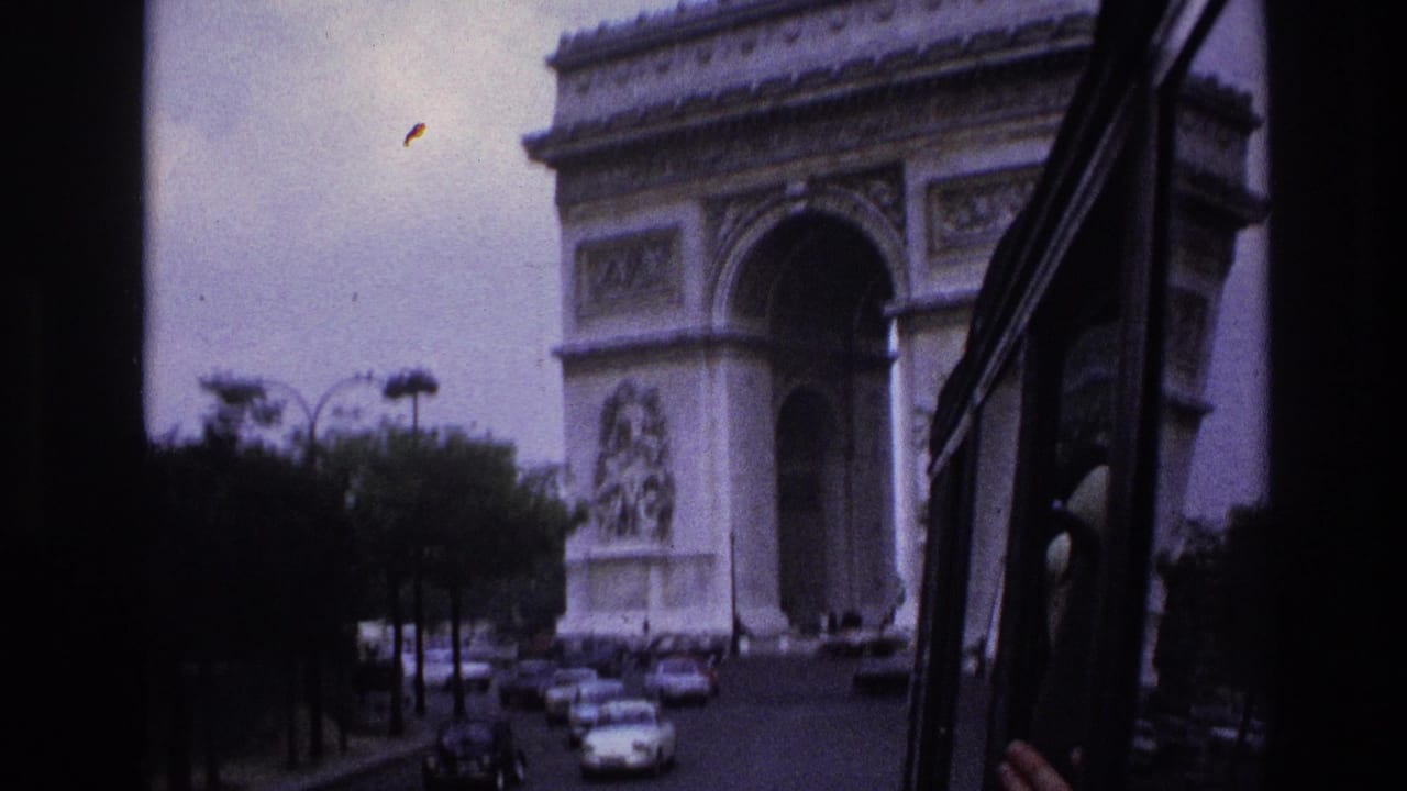 1967 Paris travel footage frame with the Arc de Triomphe ahead, seen past a dark vehicle frame and mid-century street traffic.