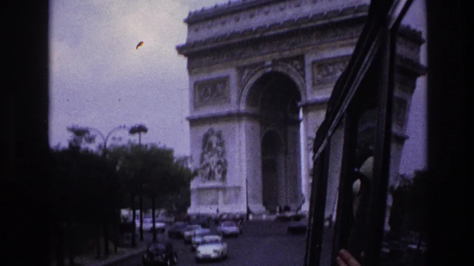 1967 Paris travel footage frame with the Arc de Triomphe ahead, seen past a dark vehicle frame and mid-century street traffic.