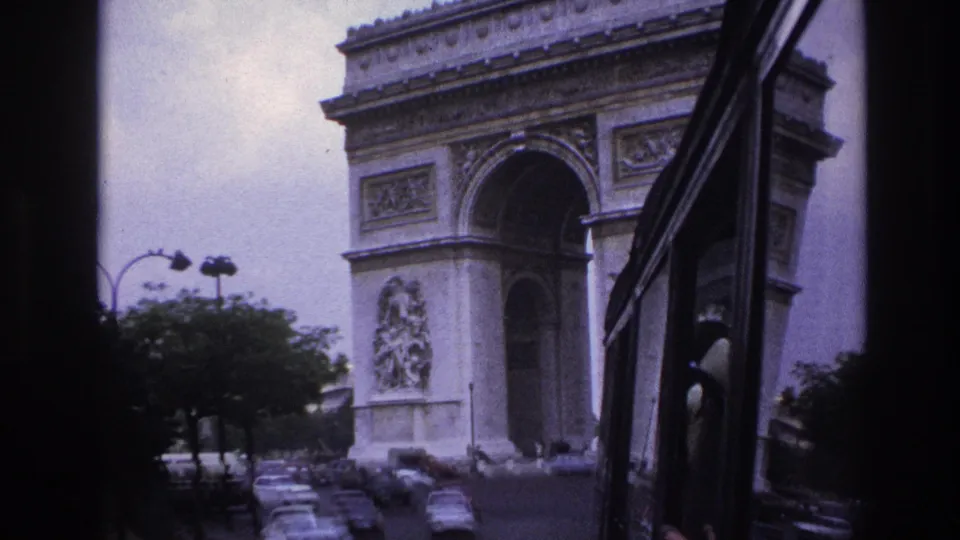 1967 Paris 8mm frame showing the Arc de Triomphe from a low moving angle, with roadway cars and a vehicle window frame at right.