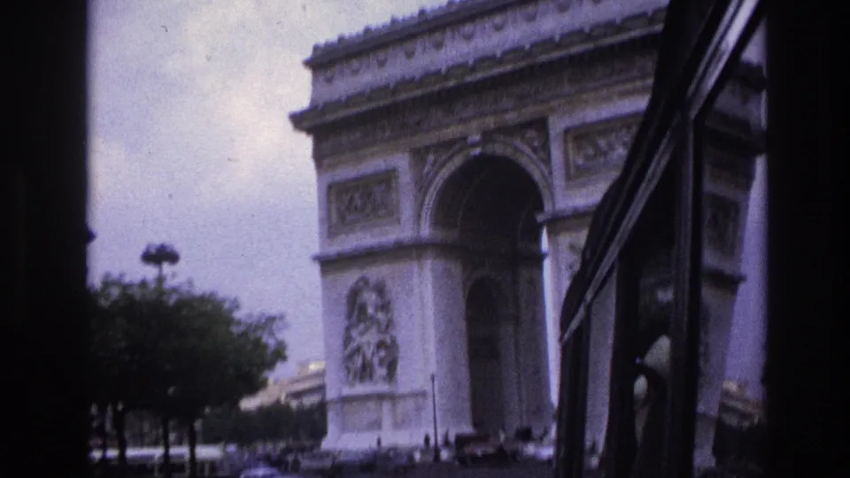1967 Paris home-movie frame of the Arc de Triomphe seen through a moving vehicle, with dark framing, trees, cars, and small pedestrians.
