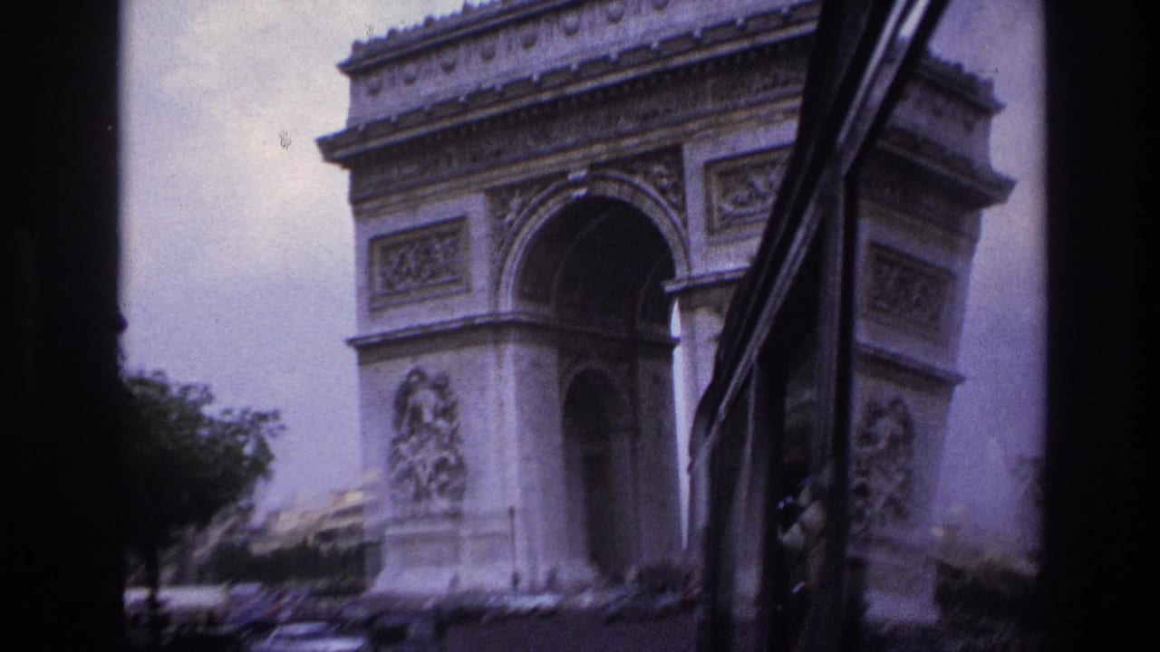 1967 Paris home-movie frame showing the Arc de Triomphe from a vehicle approach, with traffic below and dark window edges in view.