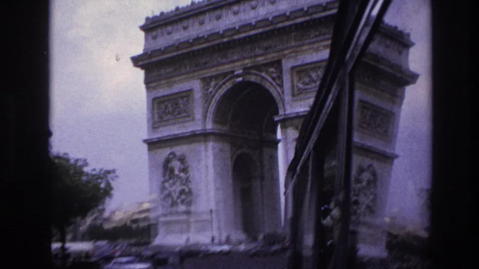 1967 Paris home-movie frame showing the Arc de Triomphe from a vehicle approach, with traffic below and dark window edges in view.