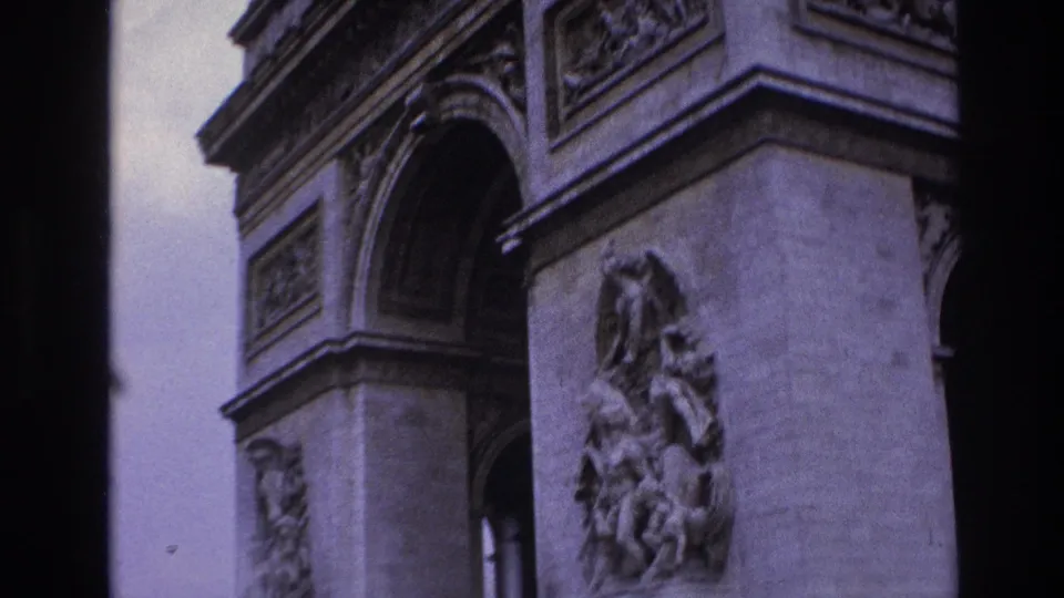 1967 Paris home-movie frame showing the Arc de Triomphe with a large sculpted relief, arch opening, carved frieze, and purple-tinted film color.