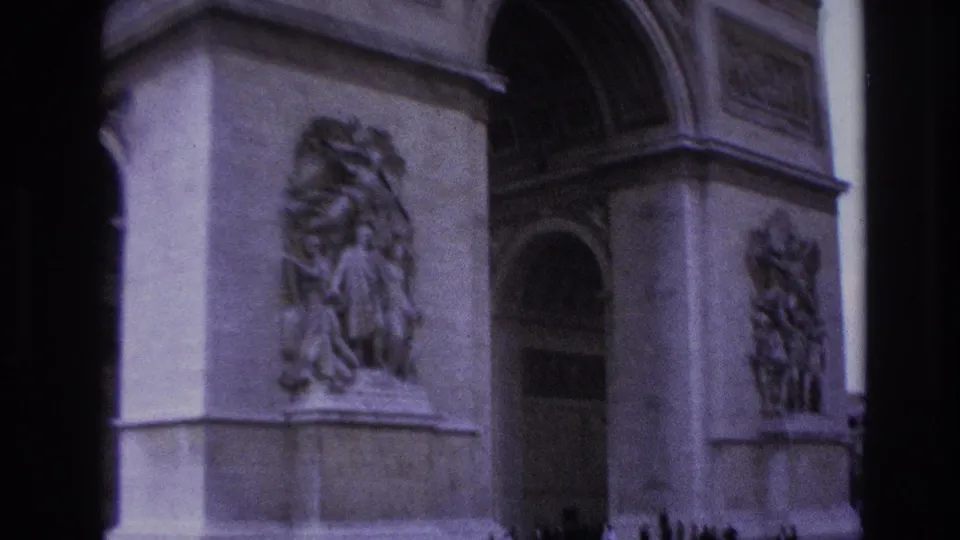 Purple-tinted 8mm frame from Paris showing two portions of the Arc de Triomphe, including sculpted reliefs, arched openings, and a dark vertical divider.
