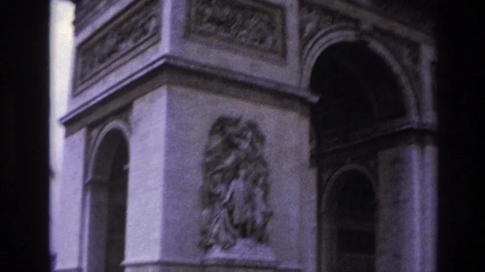 1967 home-movie frame of the Arc de Triomphe corner in Paris, with a relief sculpture, side arch, upper carving, and soft purple 8mm color.