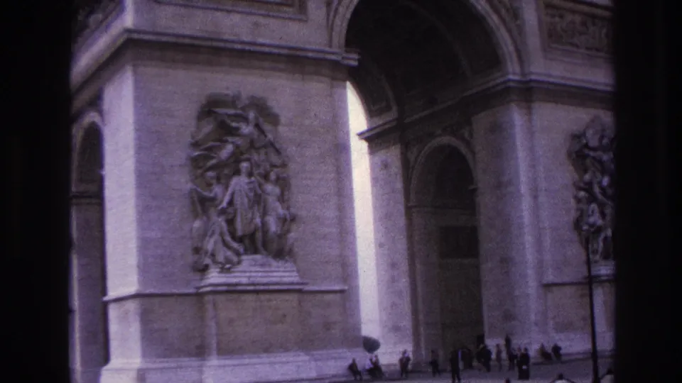 1967 Paris home-movie frame showing the Arc de Triomphe base, carved reliefs, arched openings, and small pedestrians near the monument.