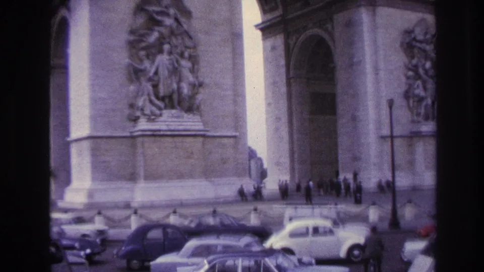 1967 Paris frame of the Arc de Triomphe seen across traffic, with pedestrians at the base and several period cars passing in front.