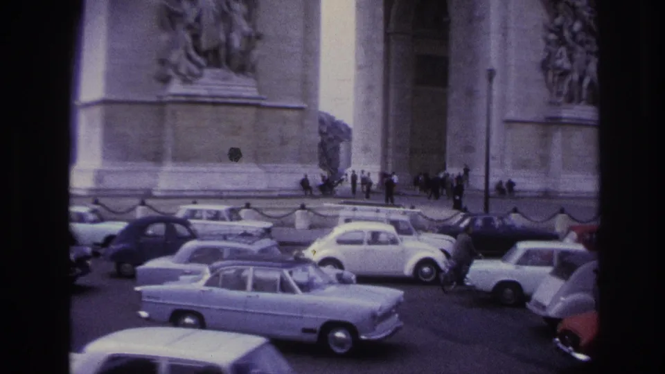 1967 Paris home-movie view of compact European cars moving near the Arc de Triomphe, with pedestrians and monument stonework behind them.