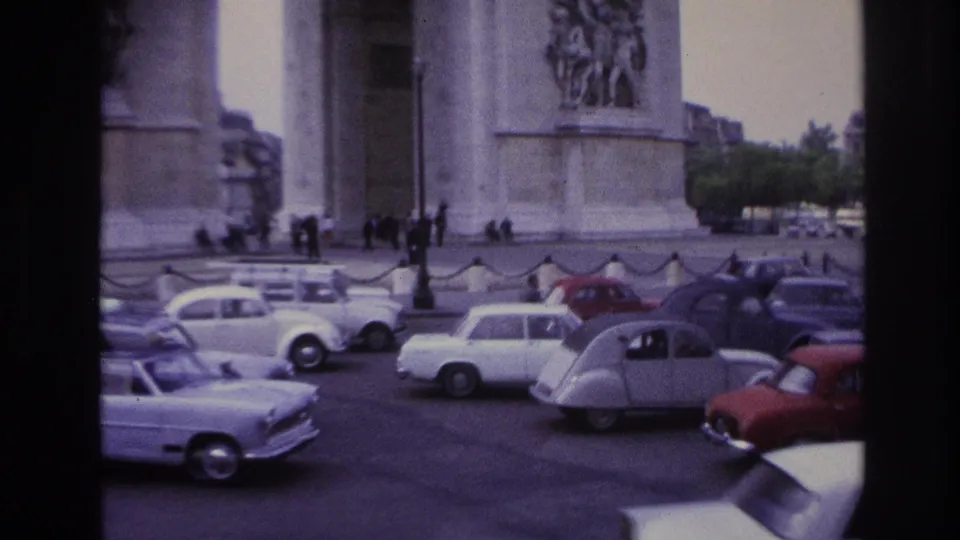 1967 Paris frame showing multiple period cars near the Arc de Triomphe, with pedestrians, sculptural reliefs, and a tree-lined edge in view.