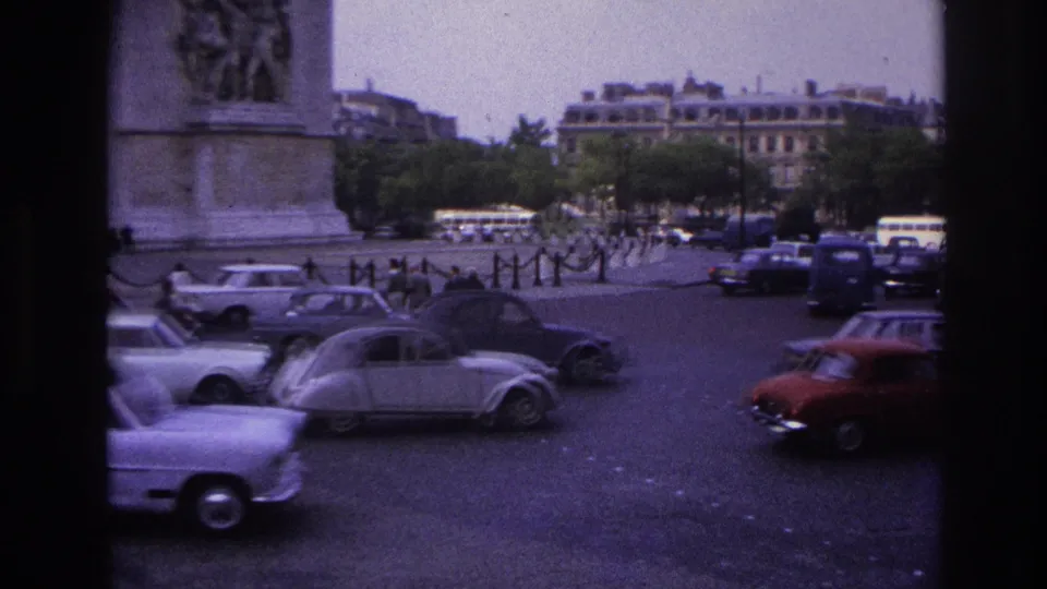 Paris traffic moves past the Arc de Triomphe, with compact sedans and vans visible in a 1960s street scene.