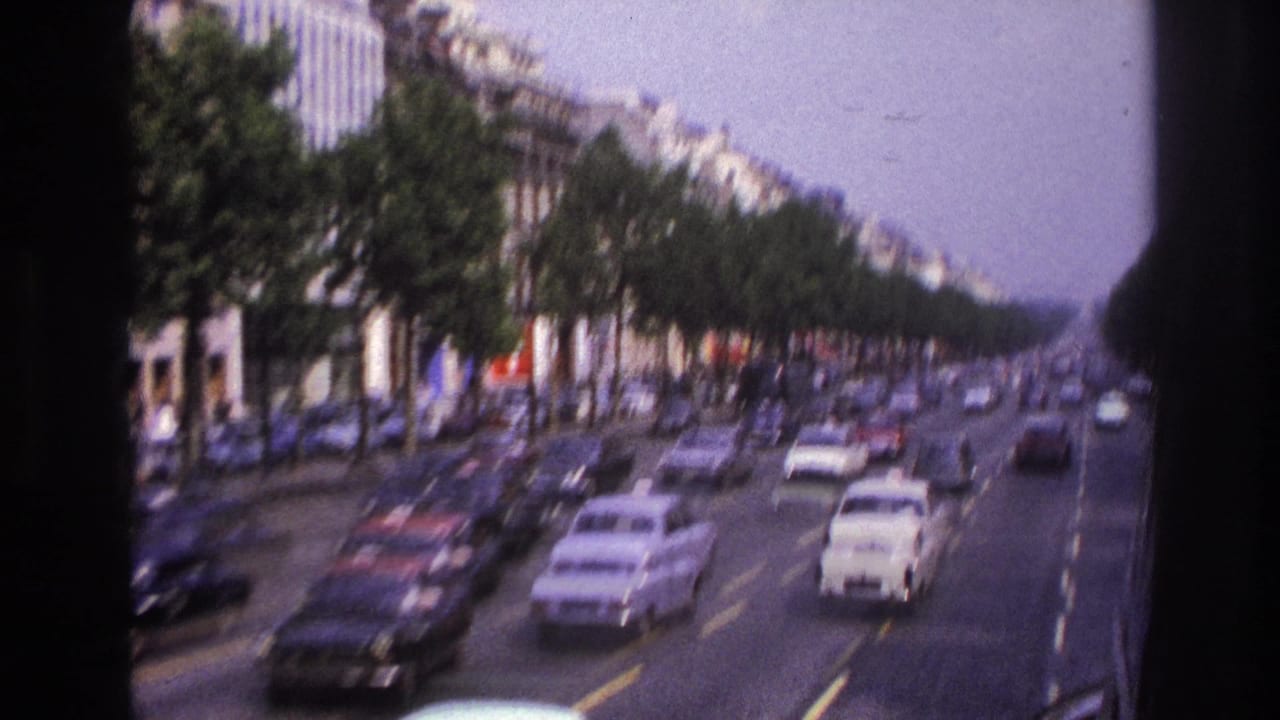 Paris boulevard traffic in 1967, with white and dark cars passing tree rows and Haussmann-style facades.