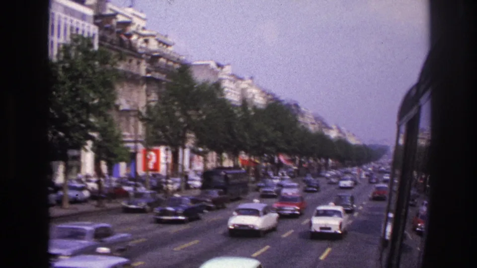 Moving bus perspective on a busy Paris street, with cars, trees, storefront color, and residential facades.