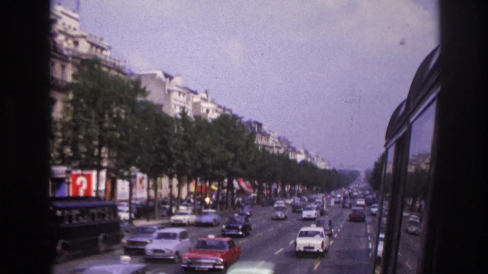 1967 Paris bus-side view over boulevard traffic, with vintage cars, tree rows, and building facades receding into the distance.