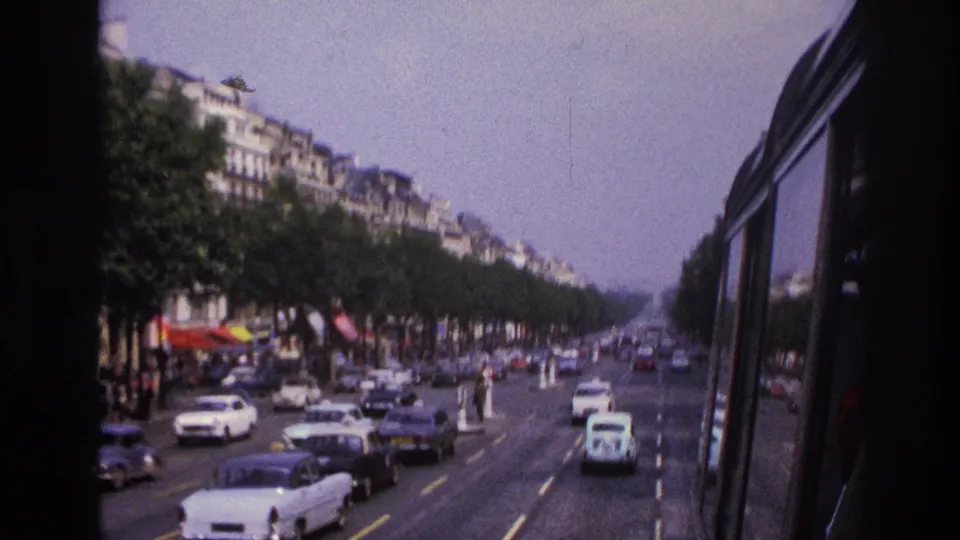 Late-1960s Paris boulevard traffic from inside a bus, with cars in several lanes, tree rows, roadside figures, and apartment facades.