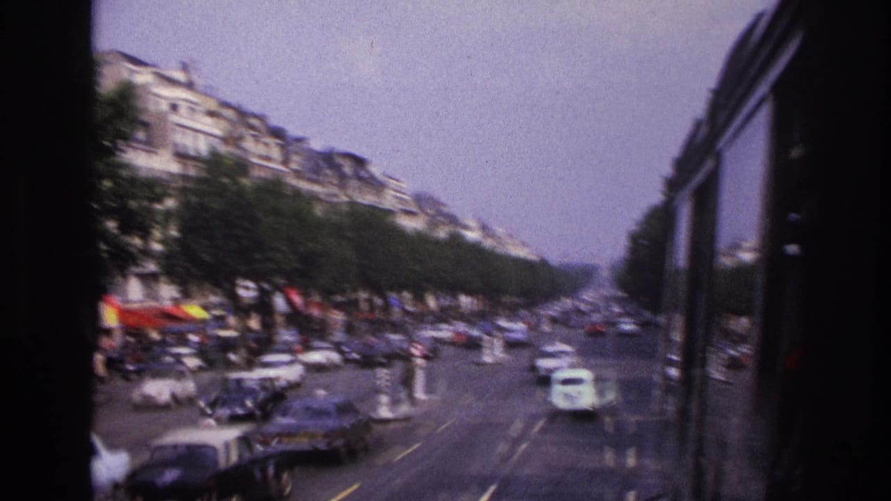 1967 Paris boulevard traffic seen from a moving vehicle, with vintage cars, trees, and apartment facades in soft 8mm focus.
