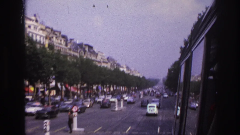 Paris street traffic in 1967, filmed through a vehicle window with roadside trees, classic cars, and distant pedestrians.