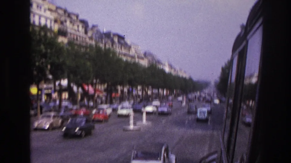 1967 Paris boulevard view with classic cars, roadside awnings, dense tree rows, and masonry buildings seen from a moving vehicle.