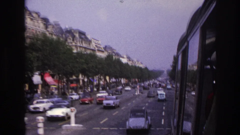 Moving-vehicle 8mm view of a 1967 Paris boulevard with vintage cars, pedestrians, trees, and apartment buildings.