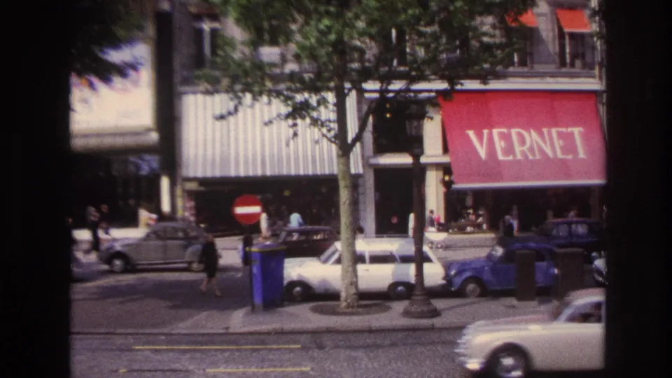 1967 Paris storefront view from a bus, showing a red VERNET awning, parked cars, pedestrians, trees, and passing traffic.