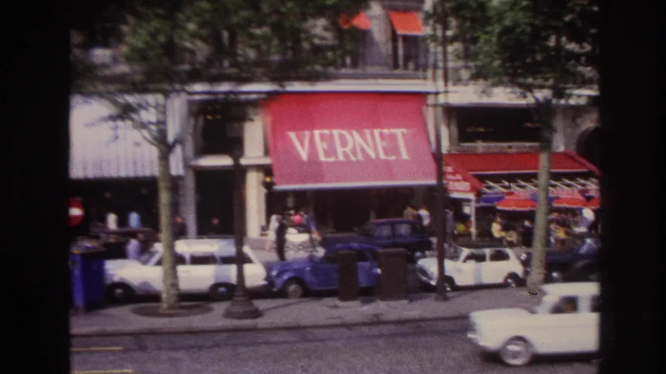 1967 Paris street-level storefronts seen from a bus, with the VERNET awning, red shop awnings, pedestrians, parked cars, and a passing white car.