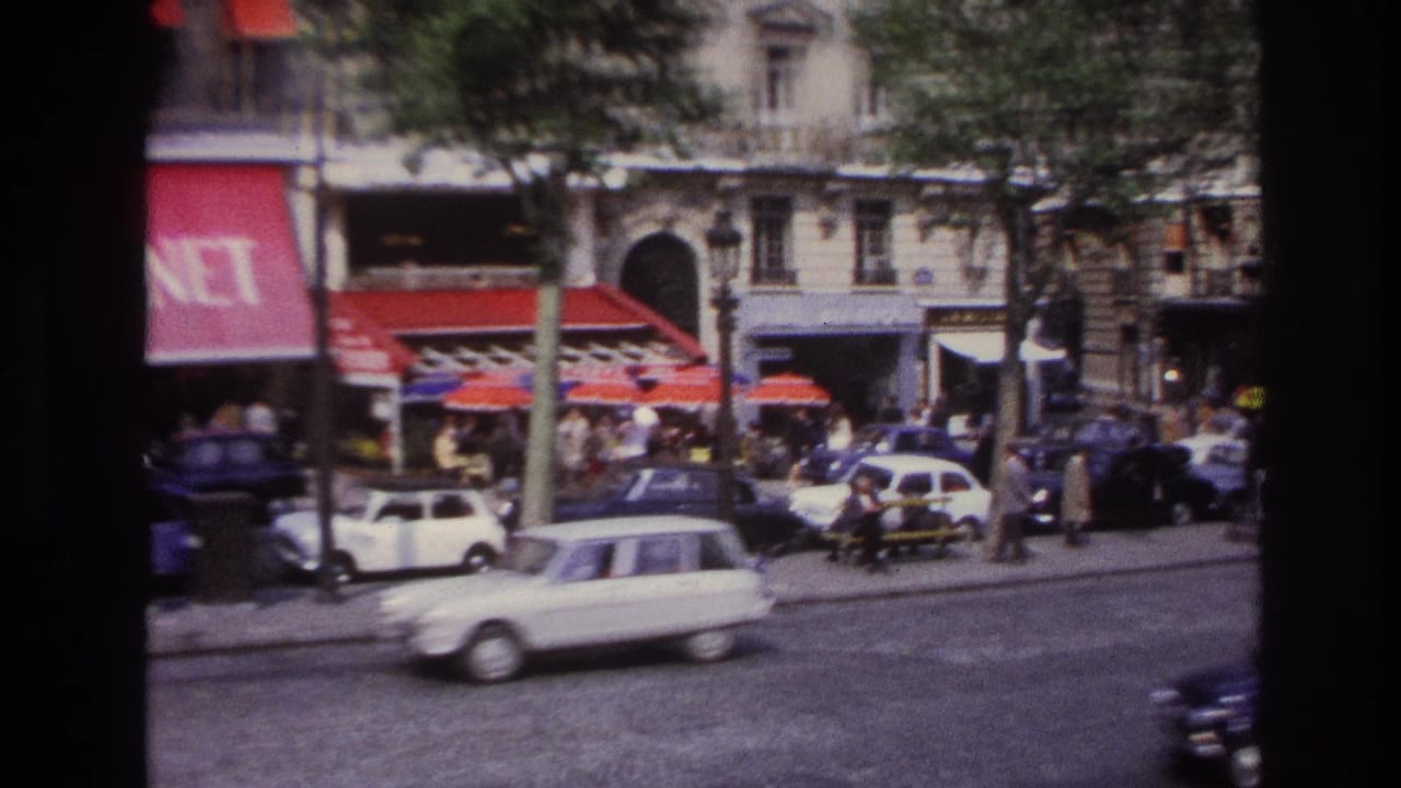 Classic cars move along a tree-lined street beside red awnings and sidewalk crowds.